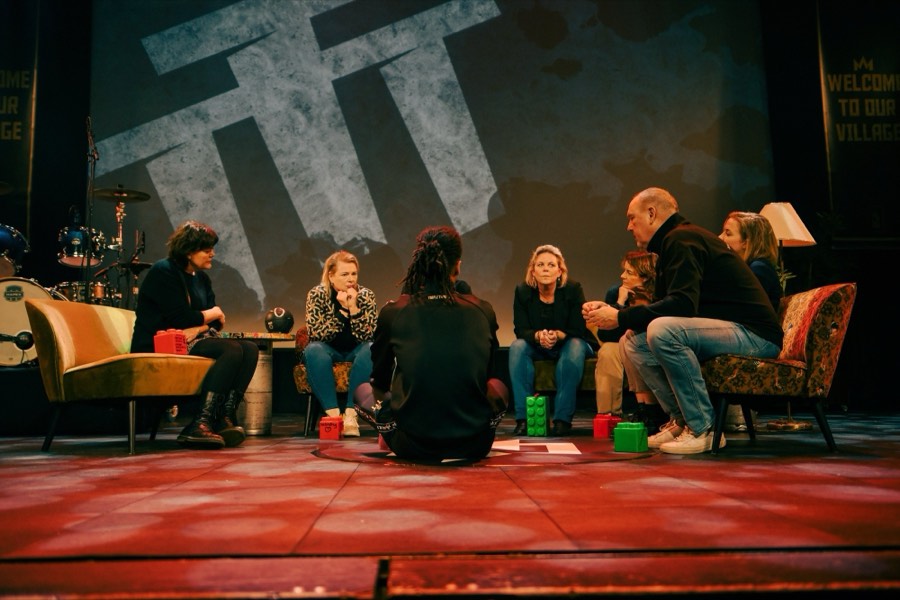 Een kring van volwassenen en een jongere in gesprek op een podium, onder het grote Triple ThreaT-logo.