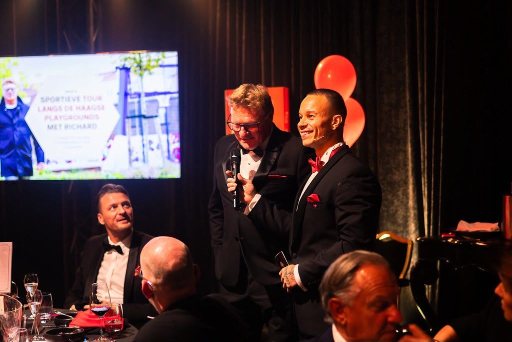 Nick Hoogendorp en een tweede presentator in smoking interviewen een gast. Op het scherm staat een tour langs de playgrounds met Richard.