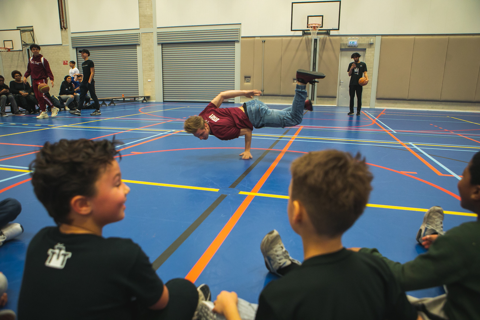 In een sporthal maakt een danser een breakdance-freeze terwijl twee jongens op de voorgrond toekijken.